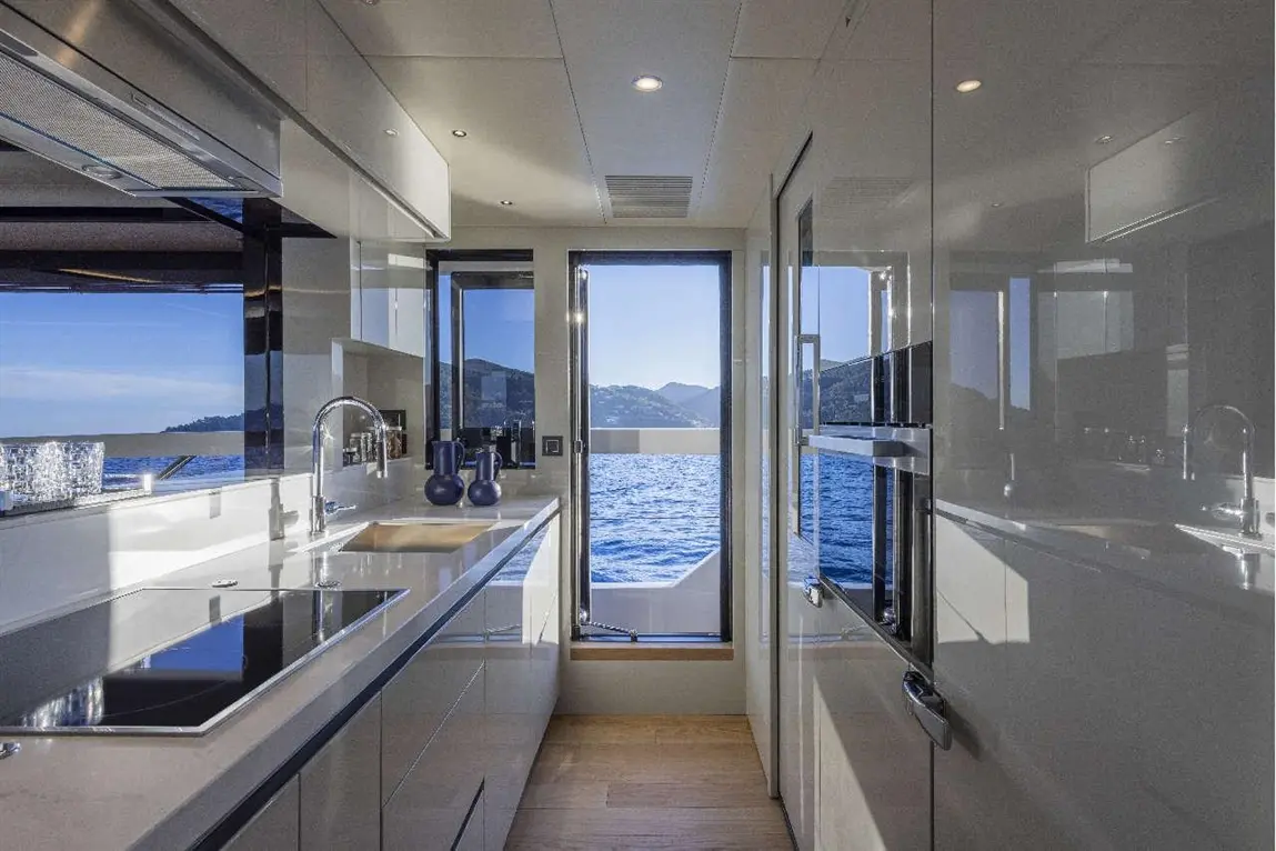 Modern yacht kitchen with sleek cabinets, a view of the sea, and mountains, featuring a stove and sink by large windows.